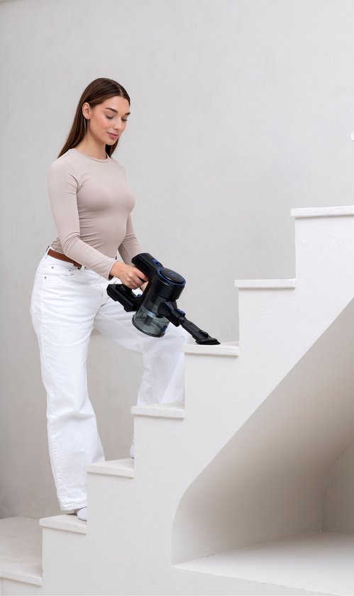 Vrouw in beige top en witte broek zuigt trap schoon met zwarte steelstofzuiger.