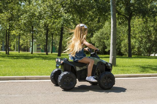 Een meisje met een witte strik in haar blonde haar, rijdend op een groene speelgoedquad met camouflageprint op een weg.