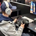 Jongeman met koptelefoon die een Thrustmaster T128 racestuur gebruikt, zittend voor een computer.
