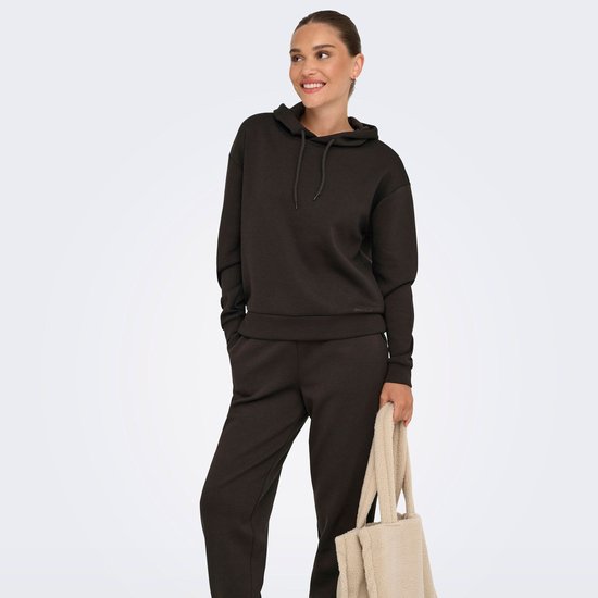 Model met een glimlach in een donkerbruine hoodie, handen in de zakken, met een beige tas in de hand.