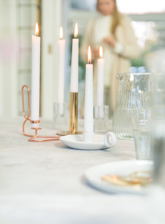 Tafel met meerdere brandende witte dinerkaarsen in koperen en gouden houders, een vrouw op de achtergrond.