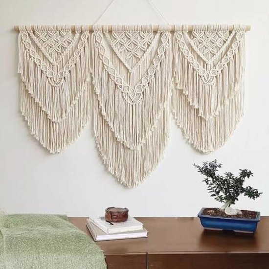 Crèmekleurig macramé wandkleed met drie V-vormige patronen, boven een bureau met boeken en een bonsai.