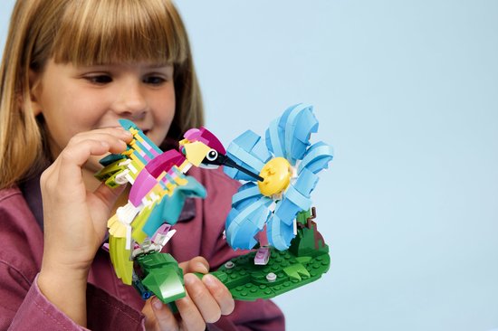 Une fille tient un colibri LEGO aux ailes vertes, jaunes et roses, près d'une fleur bleue.