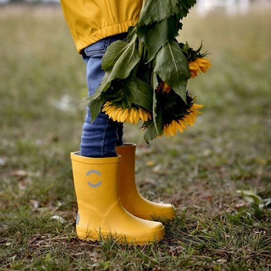 Enfant portant des bottes de pluie jaunes et une veste jaune, debout dans l'herbe avec des tournesols.
