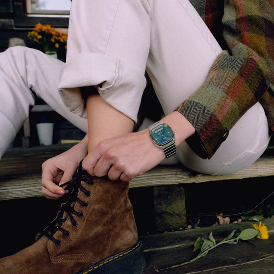 Persoon in ruitjesjas en witte broek draagt een Casio-horloge met blauwe wijzerplaat tijdens het strikken van veters.