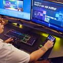 Persoon met bril speelt game, toetsenbord met RGB-verlichting, controller in handen, Hercules Stream 100 met LCD-scherm.