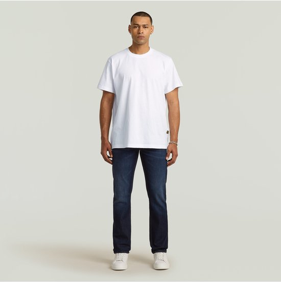 Man in een witte T-shirt en donkerblauwe G-Star jeans, staand. Volledige frontale weergave.