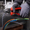 Handen met grijze handschoenen die de Knipex StepCut XL kabelschaar gebruiken om bruine, zwarte en blauwe kabels door te knippen.