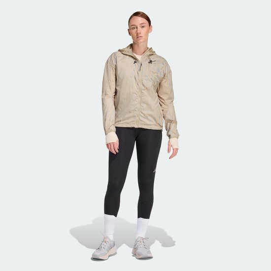 Vrouw in een beige jas met capuchon en een zwarte legging met driestrepen.