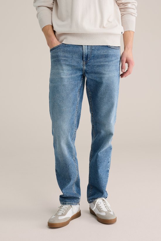 Man met de handen in de zakken, dragend een beige trui en een blauwe jeans met lichte wassing.