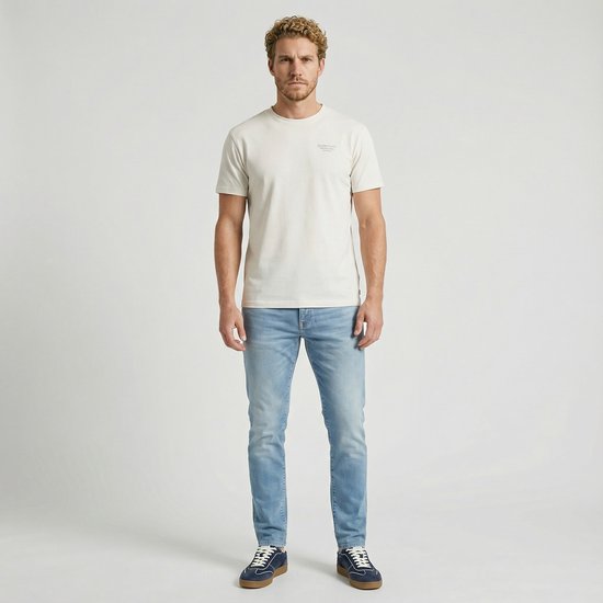 Man in een lichtblauwe jeans, wit T-shirt en blauwe sneakers. De jeans hebben een normale wassing en een slim fit pasvorm.