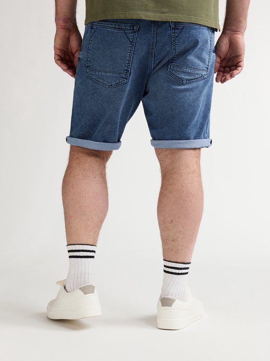 Achterkant van de onderbenen van een man die een blauwe denim short met opgerolde zoom draagt. Draagt witte sokken met zwarte strepen en witte sneakers.