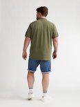 Man van achteren gezien, draagt olijfgroene polo en blauwe denim shorts met opgerolde zoom. Draagt witte sokken met zwarte strepen en witte sneakers.
