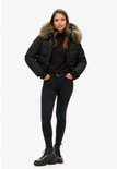 Model in een zwarte korte gewatteerde jas met bontkraag, zwarte coltrui, skinny jeans en stoere laarzen.