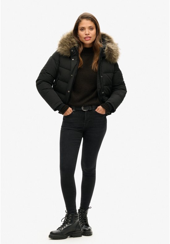 Model in een zwarte korte gewatteerde jas met bontkraag, zwarte coltrui, skinny jeans en stoere laarzen.