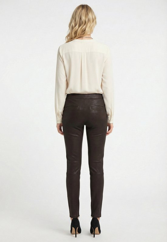 Vrouw met blond haar, achterkant, draagt een beige blouse en een donkerbruine leren broek.