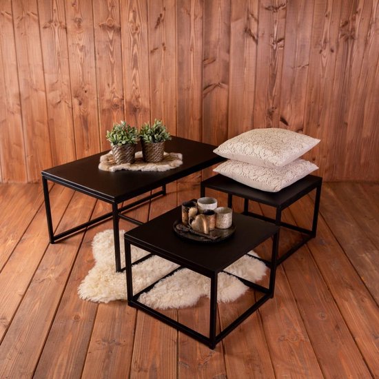 Ensemble de tables basses noires avec une grande et deux petites tables sur un plancher en bois, avec des plantes, des coussins et une peau de mouton.