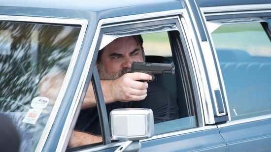 Een man met een baard en zwart shirt richt een pistool vanuit een auto.