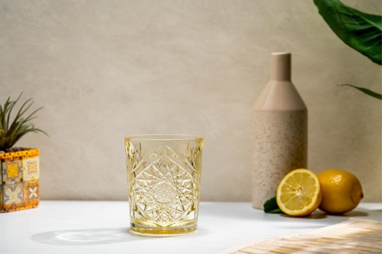 Geel, reliëf drinkglas staat op een witte tafel met citroenen en een plant.