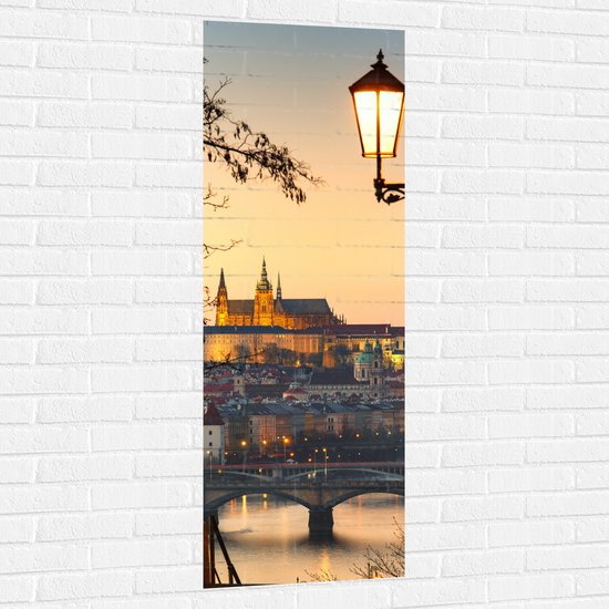 Verticale muursticker van de Praagse Burcht en een straatlantaarn bij zonsondergang op een witte bakstenen muur.