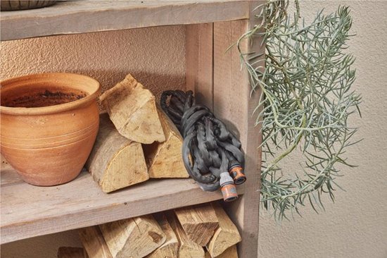 Opgerolde grijze tuinslang met oranje koppelingen op houten plank, naast pot en houtblokken.