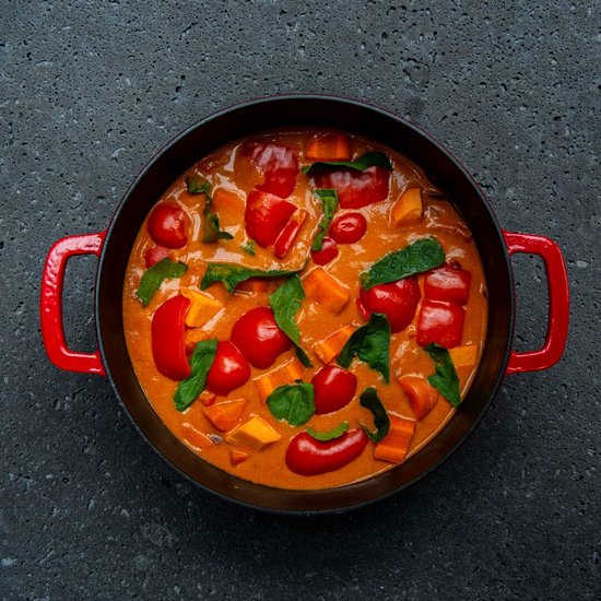 Rode gietijzeren braadpan gevuld met rode curry, zoete aardappel, paprika en spinazie, van bovenaf gezien.