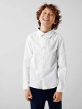 Een jongen met krullend haar draagt een wit katoenen overhemd met lange mouwen en een donkerblauwe broek.