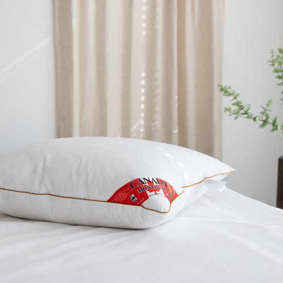 Een witte kussen met bruine bies ligt op een bed. Een rode "Canada Montreal" label en een wit label zijn zichtbaar.