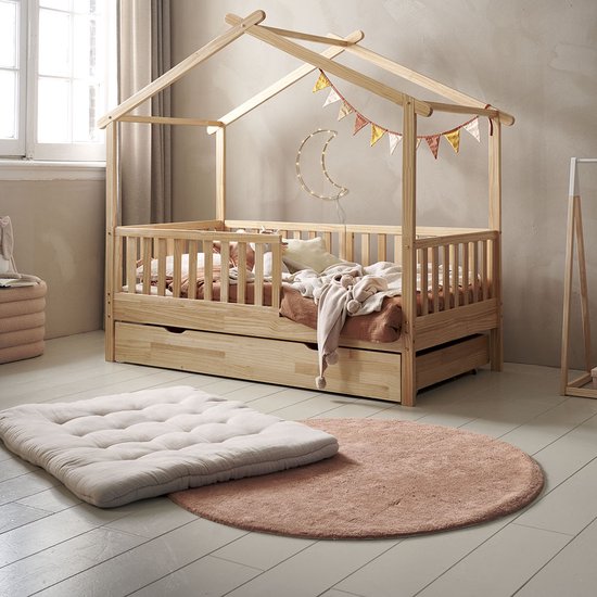 Een crème, capitonne speelkleed ligt voor een houten tipi-bed met lades, op een houten vloer met een roze vloerkleed.
