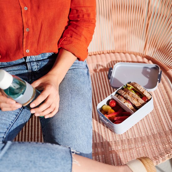 Une femme en chemise orange et jean tient une bouteille à côté d'une boîte à lunch gris clair contenant un sandwich et des légumes.