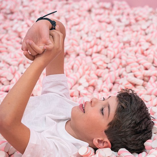 Een jongen met een one2track smartwatch om zijn pols ligt in een grote bak met roze en witte marshmallows.