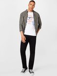 Man in een geruit overhemd over een wit T-shirt met tekst, en zwarte jeans. Draagt zwarte sneakers.