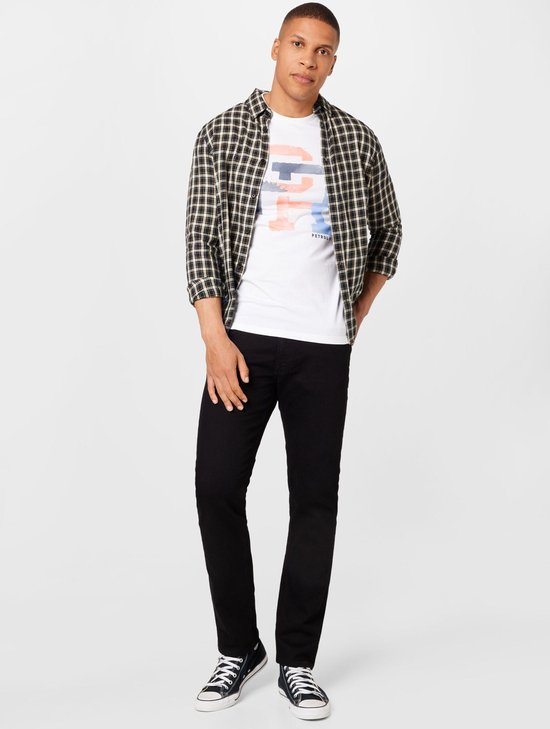 Man in een geruit overhemd over een wit T-shirt met tekst, en zwarte jeans. Draagt zwarte sneakers.