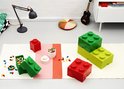 Een woonkamer met Lego opbergdozen in rood en groen, een tafellamp, een akoestisch paneel en losse Lego-blokjes op de grond.