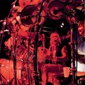 Een drummer met lang blond haar speelt op een uitgebreide drumkit, verlicht door rood licht.