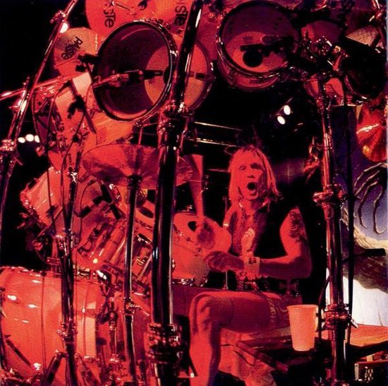 Een drummer met lang blond haar speelt op een uitgebreide drumkit, verlicht door rood licht.