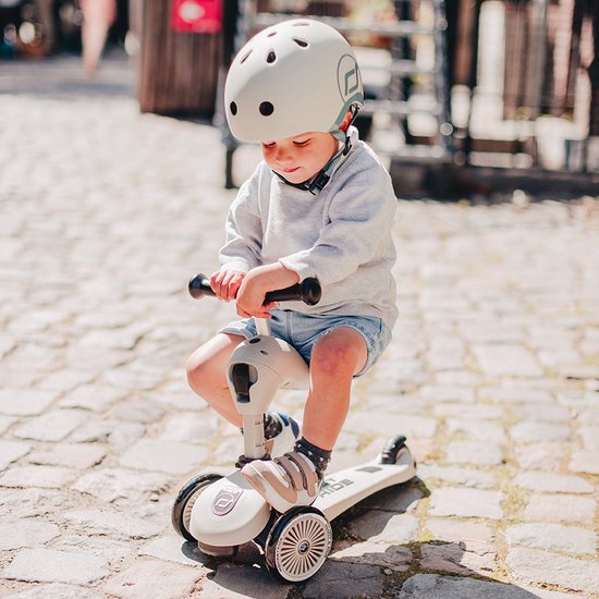 Kind op zandkleurige loopfiets met drie wielen. Tekst "SCOOT AND RIDE" op het frame.