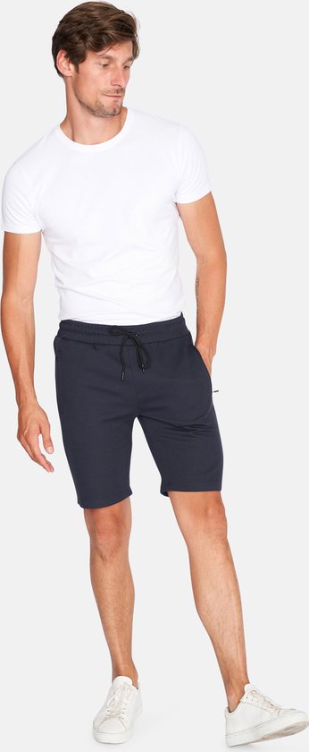 Man met witte t-shirt en donkerblauwe korte broek met trekkoord, staand en hand in de zak.
