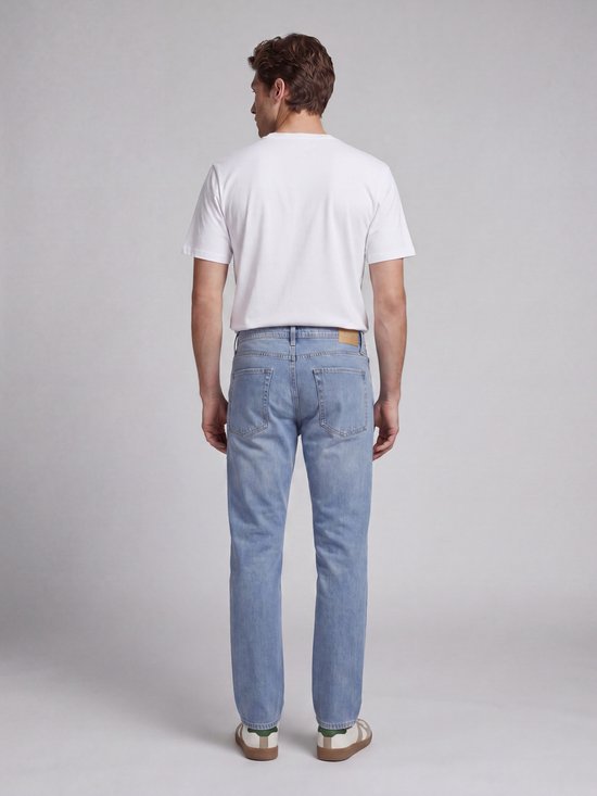 Man met witte t-shirt en lichtblauwe, rechte jeans met bruin label. Achteraanzicht.