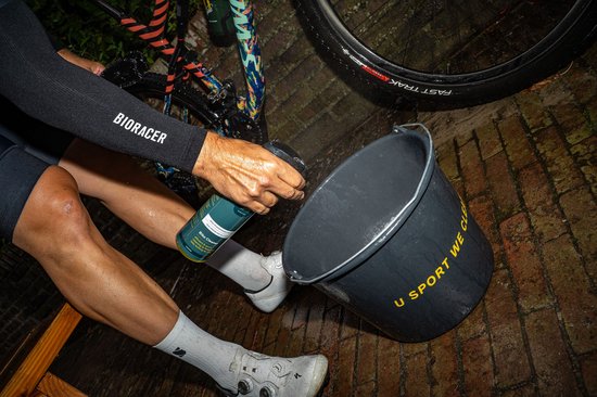 Persoon spuit U-Sport Bike Cleaner in een emmer. Fles groen met geel, emmer zwart met gele tekst "U SPORT WE CLEAN".