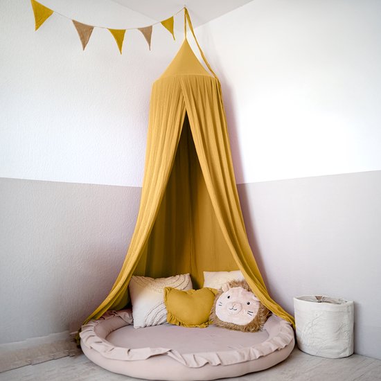 Okergele mousseline klamboe met gouden accenten en bijpassend rond speelkleed met leeuwenkussen in kinderkamer.