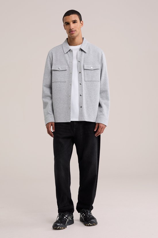 Grijs fijngebreid overshirt met knoopsluiting en borstzakken, gecombineerd met een wit T-shirt en zwarte broek.