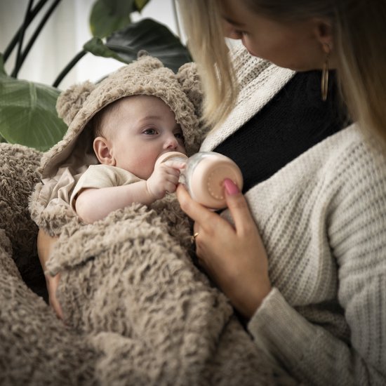 Un bébé dans une couverture enveloppante marron avec des oreilles d'ours est nourri au biberon.