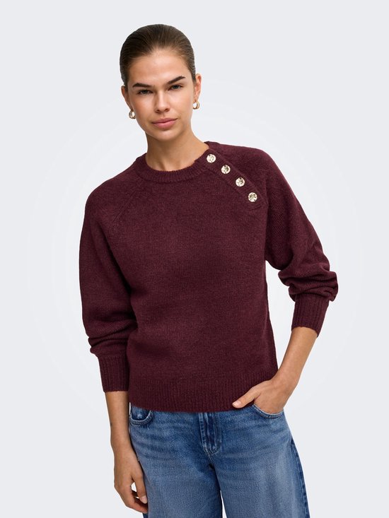 Model met een wijnrode gebreide trui met gouden knopen op de schouder en jeans.