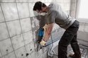 Man in werkkleding met masker boort gaten in een betegelde muur met een blauwe Bosch boorhamer.