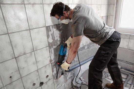 Man in werkkleding met masker boort gaten in een betegelde muur met een blauwe Bosch boorhamer.