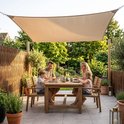 Familie eet aan een houten tafel onder een beige vierkant schaduwdoek, omringd door groen.