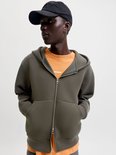 Model draagt een olijfgroen vest met rits over een oranje shirt met 'jcore' logo en een zwarte pet.