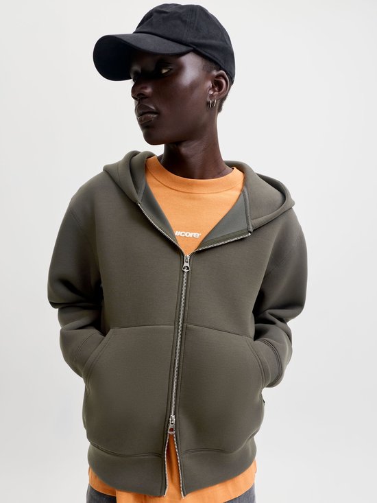 Model draagt een olijfgroen vest met rits over een oranje shirt met 'jcore' logo en een zwarte pet.