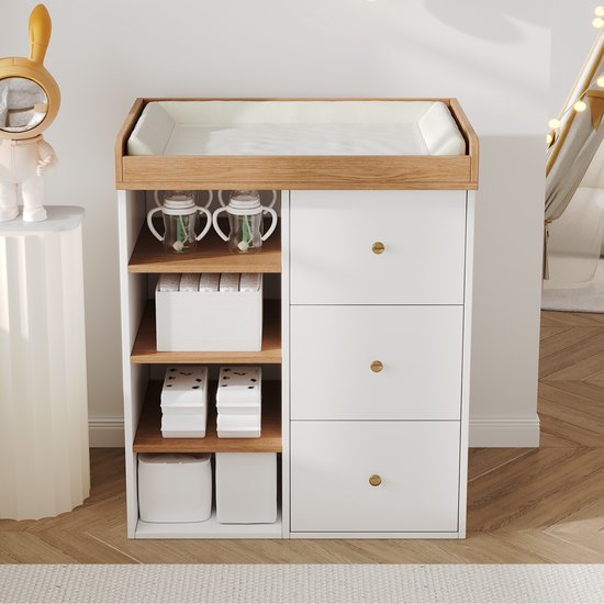 Wit-houten babycommode met aankleedkussen, drie lades met gouden knoppen en open zijplanken met opbergdozen.